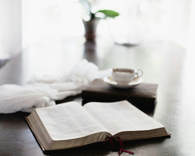 An open Bible and a cup on a table beside a bright window.