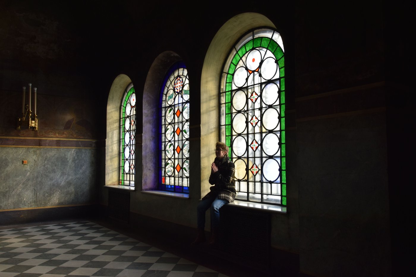 A person sitting with hands joined in prayer beside three stained-glass windows.